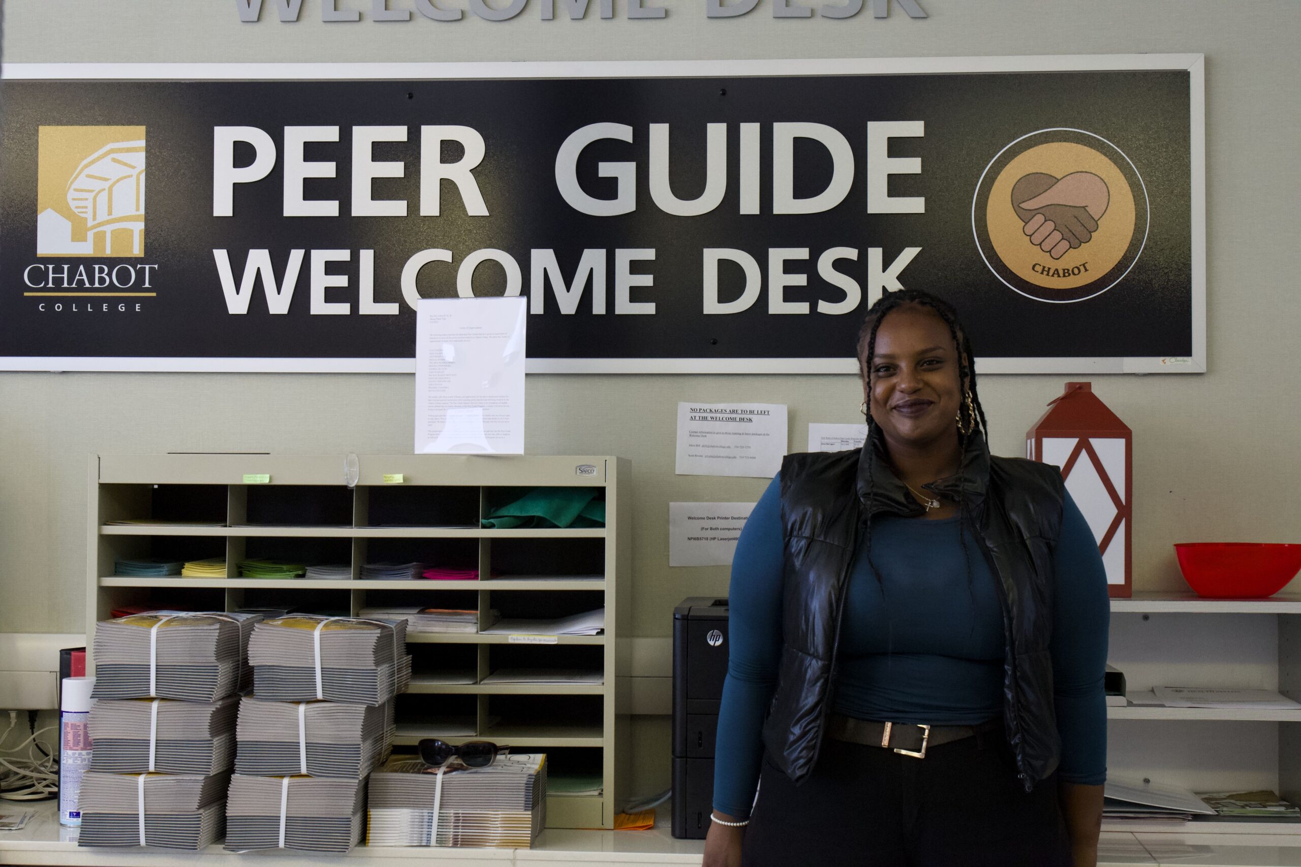 An African young woman stands in front of the Peer Guide Welcome Desk sign in Building 700.